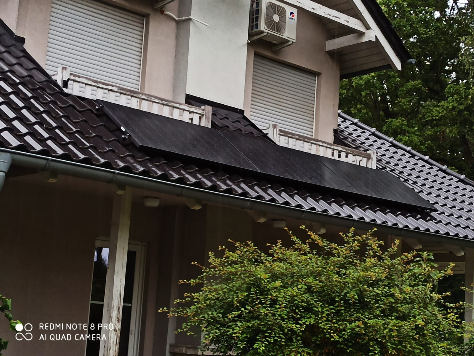 Część dachu domu jednorodzinnego z zamontowanymi panelami słonecznymi w deszczowy dzień, widoczne okna z roletami i klimatyzator na elewacji.