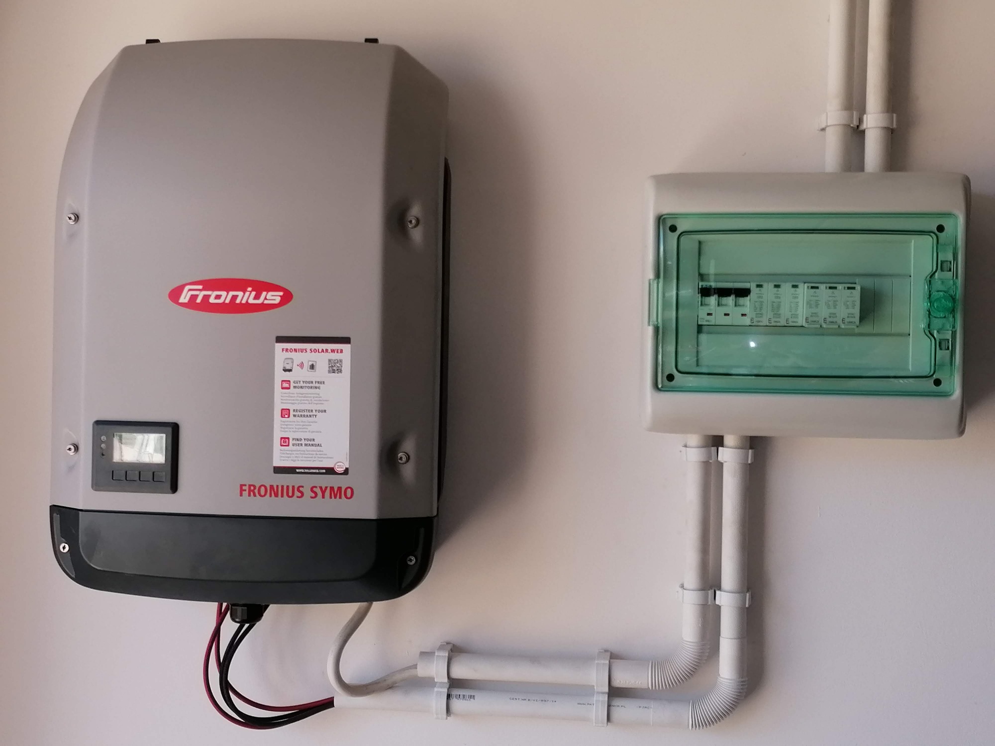 Szary inwerter Fronius Symo zamontowany na białej ścianie, obok skrzynka bezpiecznikowa z zieloną pokrywą i białymi rurami instalacyjnymi.