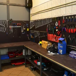 Warsztat samochodowy: ściana z narzędziami (klucze, kombinerki) nad stołem roboczym z częściami zamiennymi i pojemnikami na oleje.