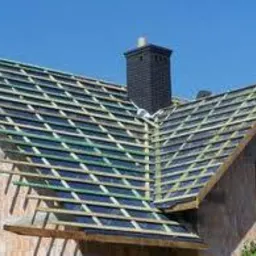 Konstrukcja dachu w trakcie budowy, widoczne krokwie, membrana dachowa, komin z cegły klinkierowej na tle błękitnego nieba.