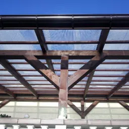 Drewniana konstrukcja z przezroczystym zadaszeniem, widok od dołu, brązowe rynny na górze, błękitne niebo w tle.