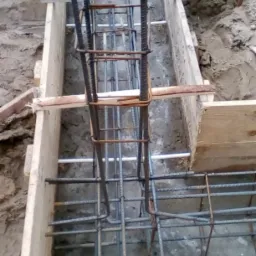 Zbrojenie pionowe i poziome w szalunku fundamentowym, przygotowane do zalania betonem; widoczne stalowe pręty zbrojeniowe i drewniany szalunek.