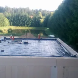 Fundamenty domu murowanego w Grudusku, zabezpieczone folią, z widokiem na pobliski staw i zalesiony teren.