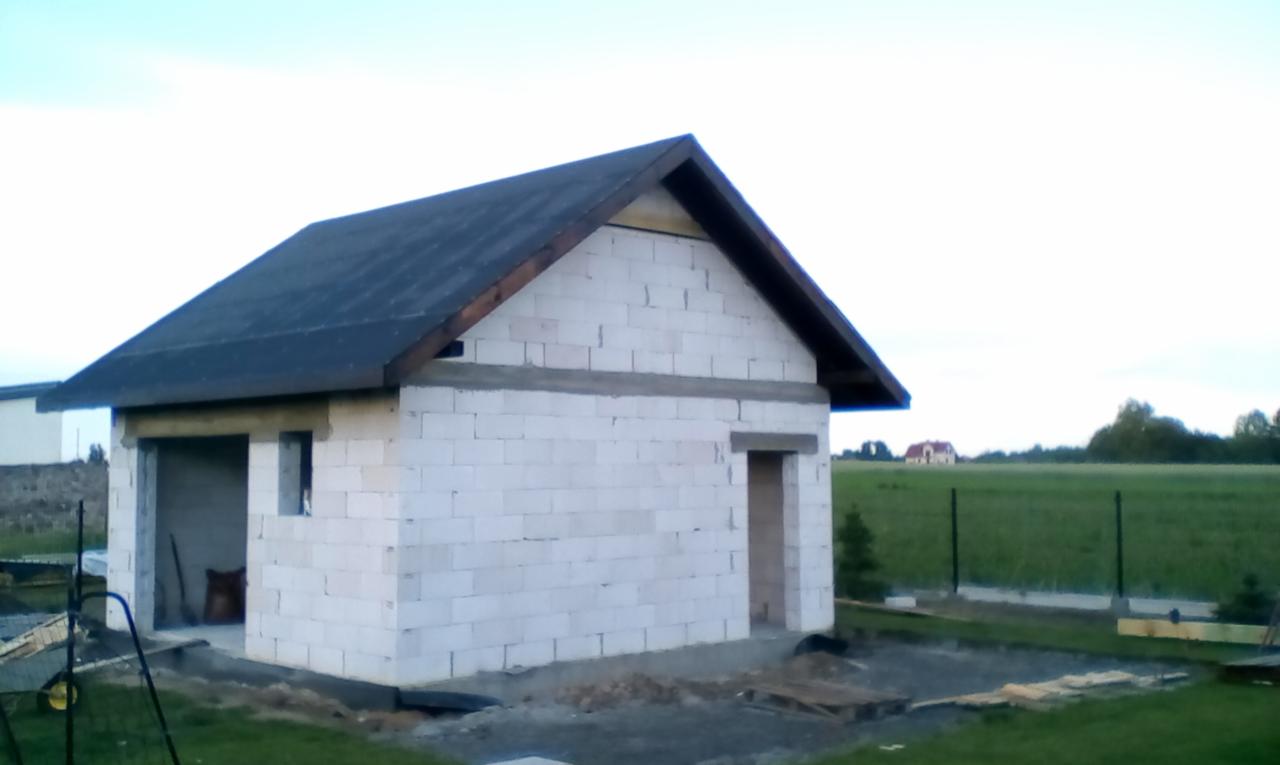 Niedokończony budynek z białych bloczków z dachem z papy, widoczne otwory okienne i drzwiowe, narzędzia wewnątrz garażu, krajobraz polny w tle.