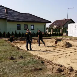Przygotowanie terenu pod budowę domu jednorodzinnego: wyrównany piasek, widoczne ślady po zagęszczarce, pracownicy w czarnych koszulkach z logo firmy budowlanej, w tle dom i materiały budowlane.