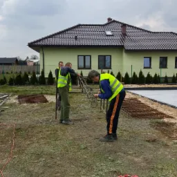 Ekipa budowlana w kamizelkach odblaskowych montuje zbrojenie na przygotowanym fundamencie, widoczne siatki zbrojeniowe, w tle dom jednorodzinny i kamera na statywie.