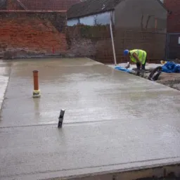 Świeżo wylana, mokra wylewka betonowa na budowie, z widocznymi rurami i narzędziami, pracownik w odblaskowej kamizelce wygładza powierzchnię.