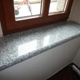 Szary, polerowany parapet z kamienia o zaokrąglonej krawędzi, zamontowany pod drewnianym oknem. W szybie okna odbija się rower.