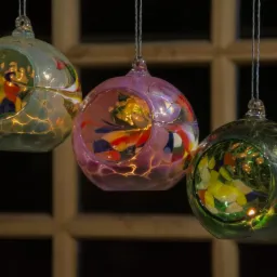 Trzy szklane kule w odcieniach zieleni, różu i błękitu, wiszące na cienkich sznurkach, wypełnione kolorowymi szkiełkami i podświetlone od wewnątrz, na tle okna z ciemnym tłem.