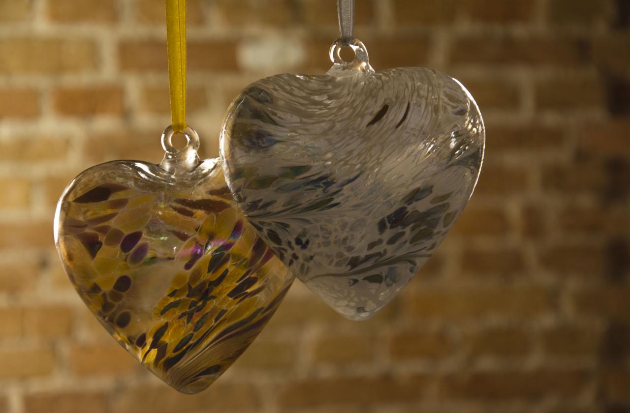 Dwa szklane serca z kolorowymi inkluzjami, zawieszone na wstążkach, na tle ceglanej ściany.