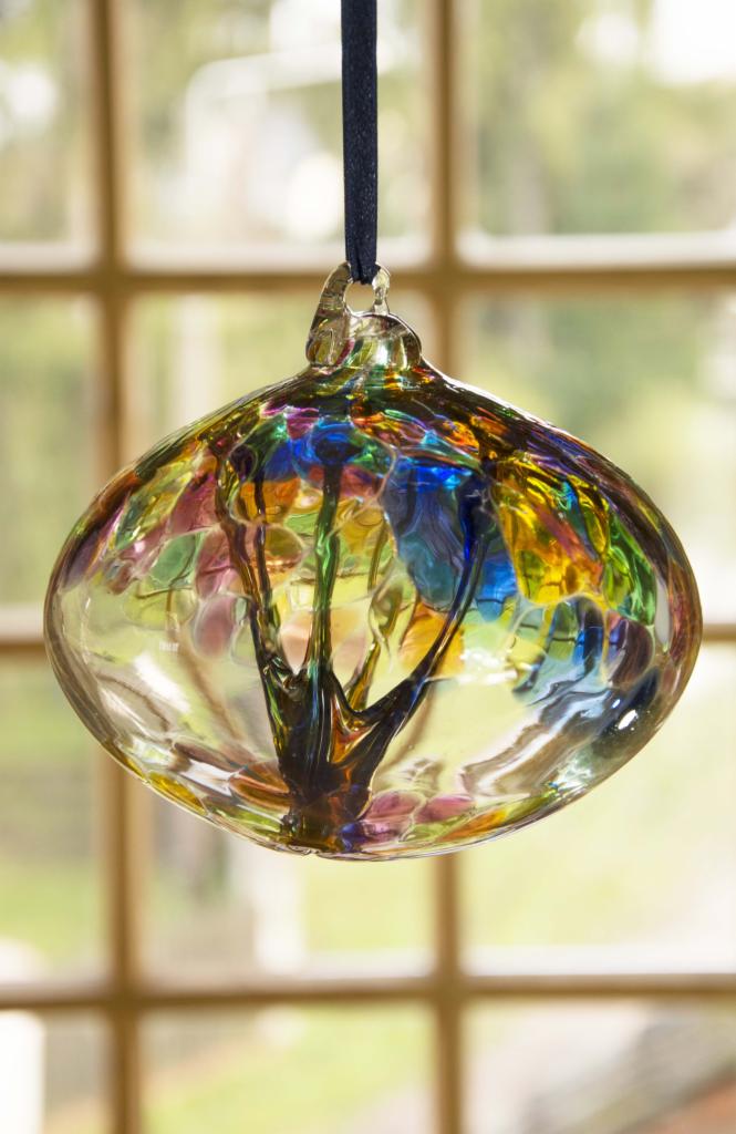 Ręcznie dmuchana, szklana kula dekoracyjna z wielobarwnymi plamkami i ciemnym wzorem przypominającym drzewo, zawieszona na czarnym sznurku na tle okna.