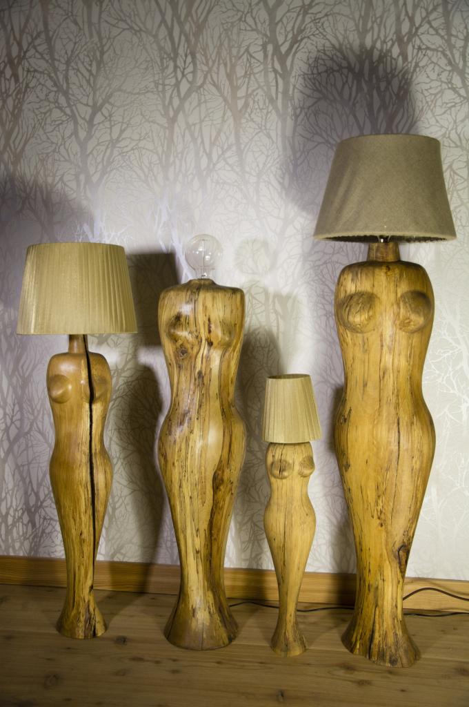 Cztery lampy stojące, każda wyrzeźbiona z drewna w kształcie kobiecej sylwetki, z abażurami w różnych odcieniach beżu, ustawione na drewnianej podłodze na tle tapety w delikatne wzory drzew.