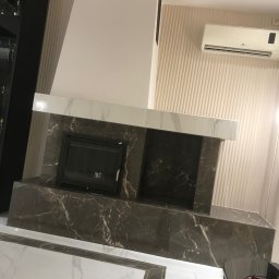 DM Design Damian Mielczarek - Wykończony kominek z czarnego marmuru z białymi żyłkami w salonie w Piotrkowie Trybunalskim, widoczna klimatyzacja na ścianie z dekoracyjnymi pionowymi panelami.