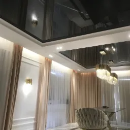 Nowoczesne wnętrze z czarnym, błyszczącym sufitem napinanym, odbijającym kryształowy żyrandol i złote kinkiety, jasne zasłony, białe ściany z dekoracyjnymi listwami i marmurowa podłoga.