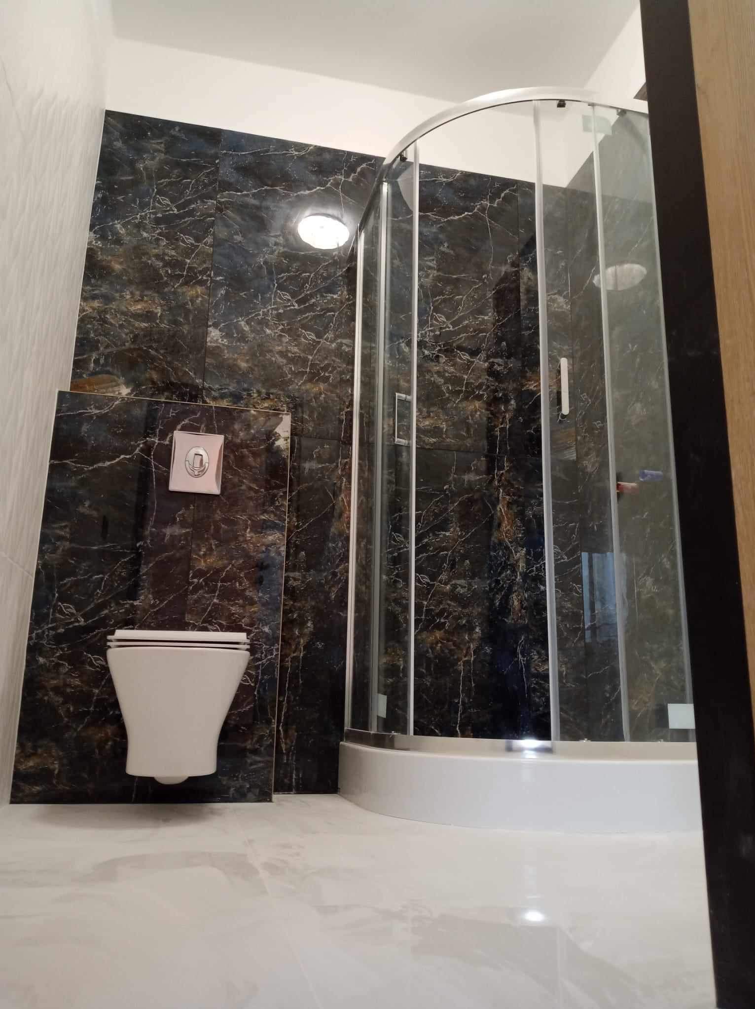 Nowoczesna łazienka z ciemnymi płytkami imitującymi marmur, kabina prysznicowa z zaokrąglonymi drzwiami i podwieszana toaleta z chromowanym przyciskiem spłukiwania.