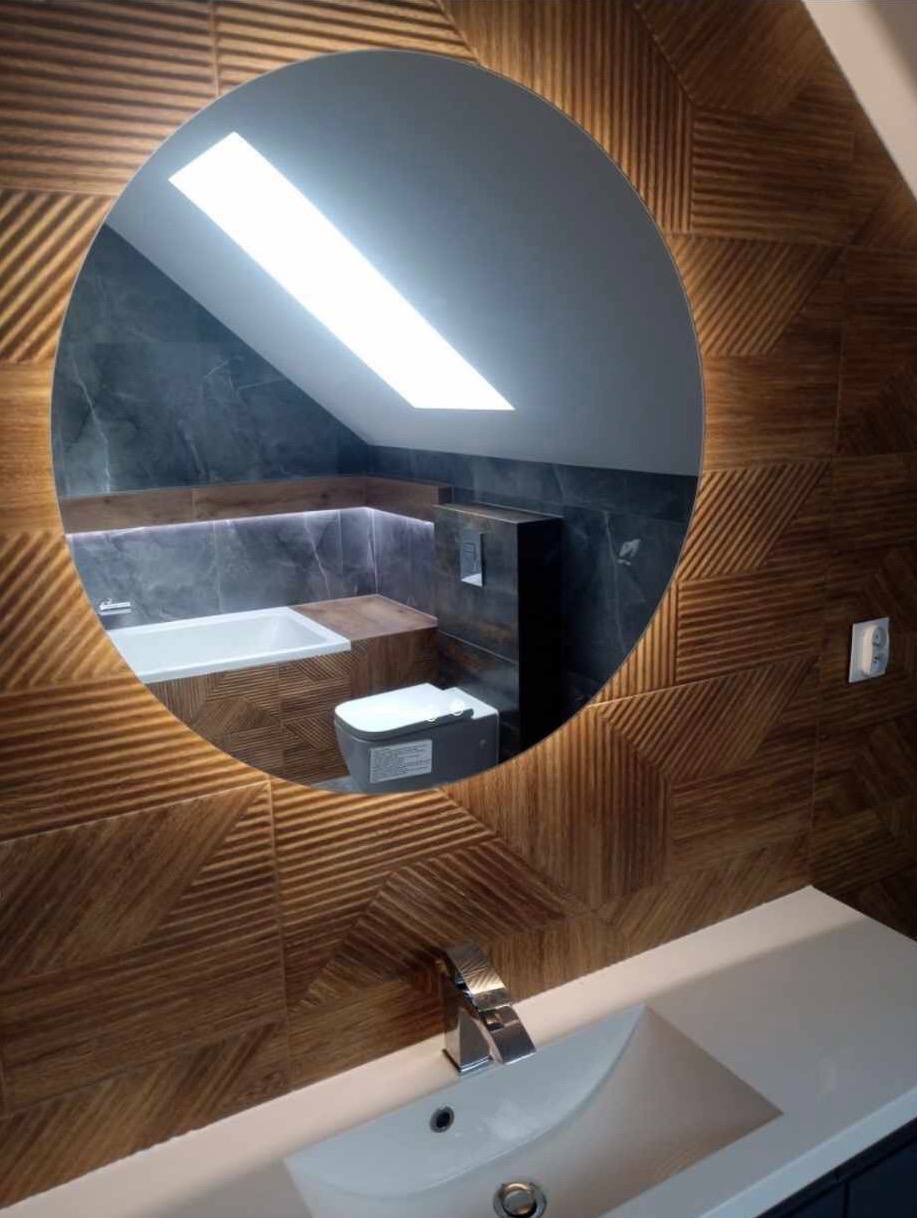 Okrągłe lustro z podświetleniem LED odbija wnętrze łazienki na poddaszu: wanna, toaleta podtynkowa, szare płytki i skośny sufit z oknem dachowym.