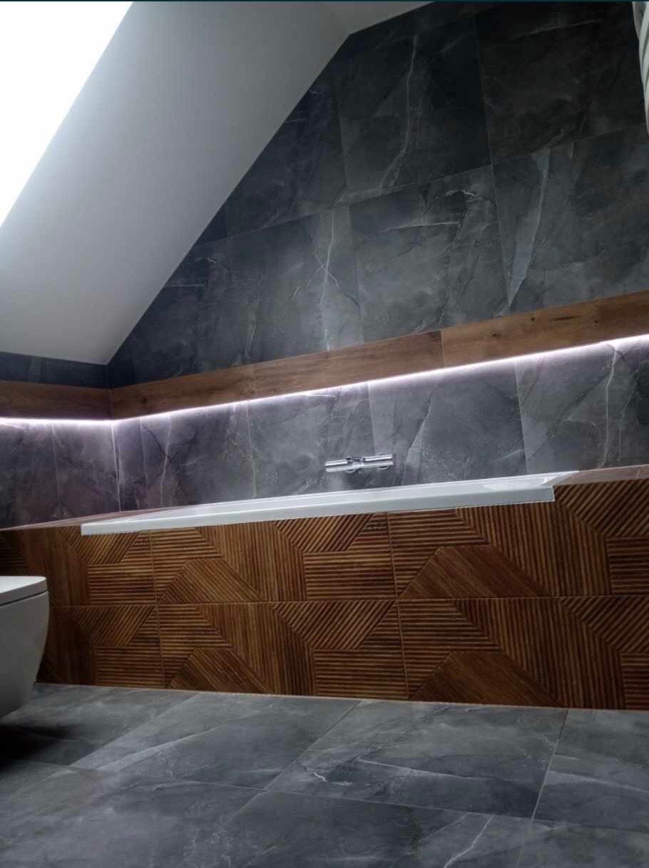 Nowoczesna łazienka na poddaszu: szare płytki imitujące marmur na ścianach, drewnopodobne panele z geometrycznym wzorem przy wannie, podświetlenie LED, szara podłoga i fragment toalety.