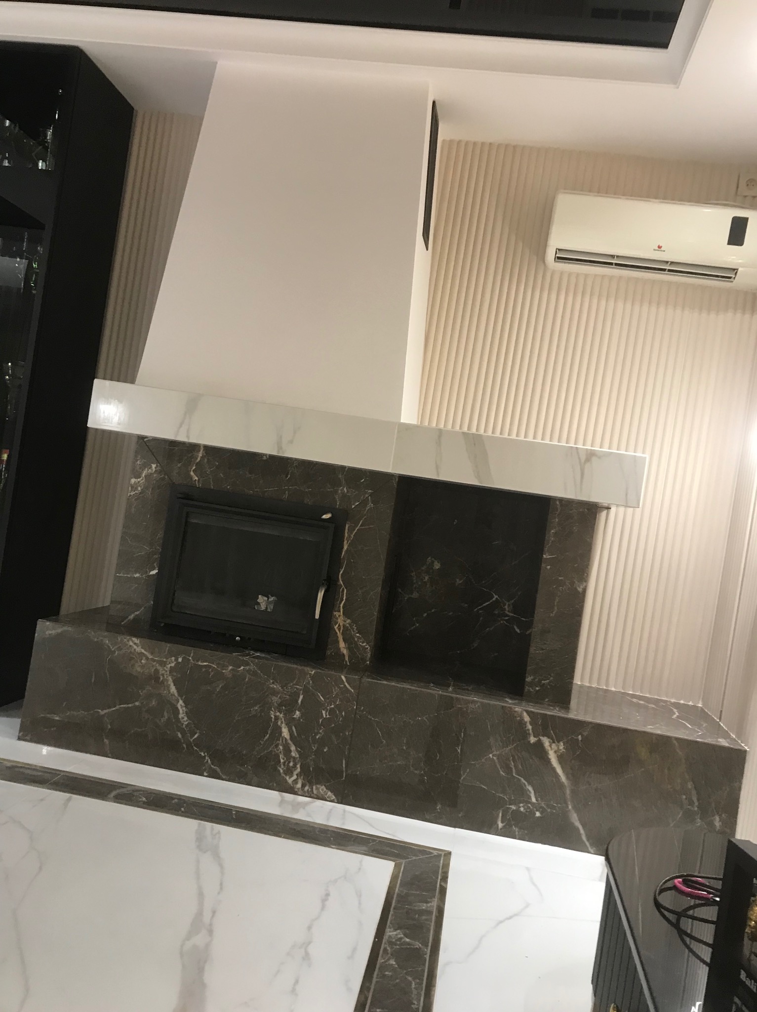 Wykończony kominek z czarnego marmuru z białymi żyłkami w salonie w Piotrkowie Trybunalskim, widoczna klimatyzacja na ścianie z dekoracyjnymi pionowymi panelami.