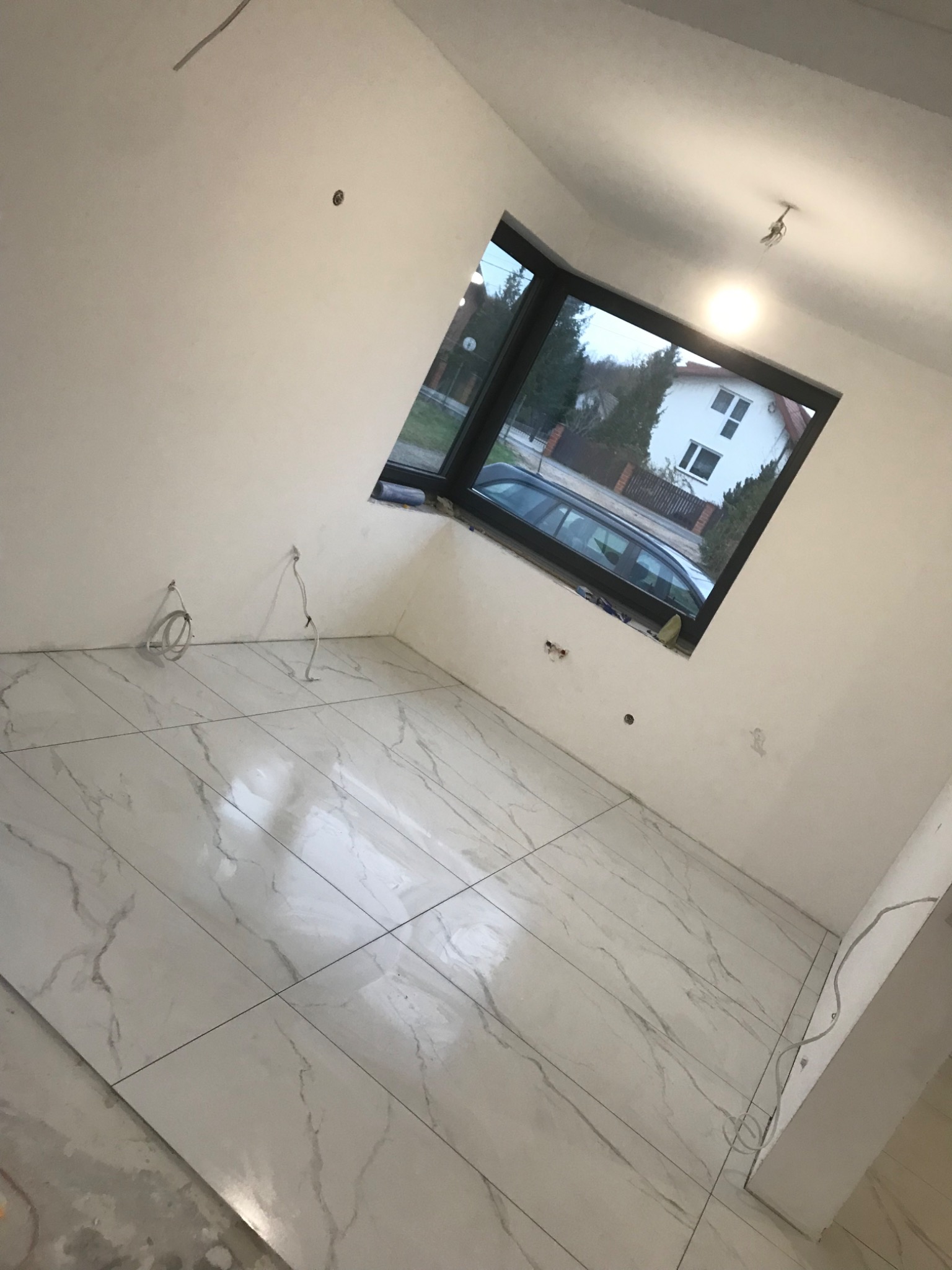 Wnętrze pokoju z ułożonymi białymi płytkami podłogowymi imitującymi marmur, widoczne wystające przewody elektryczne ze ścian, okno z widokiem na dom i samochód.