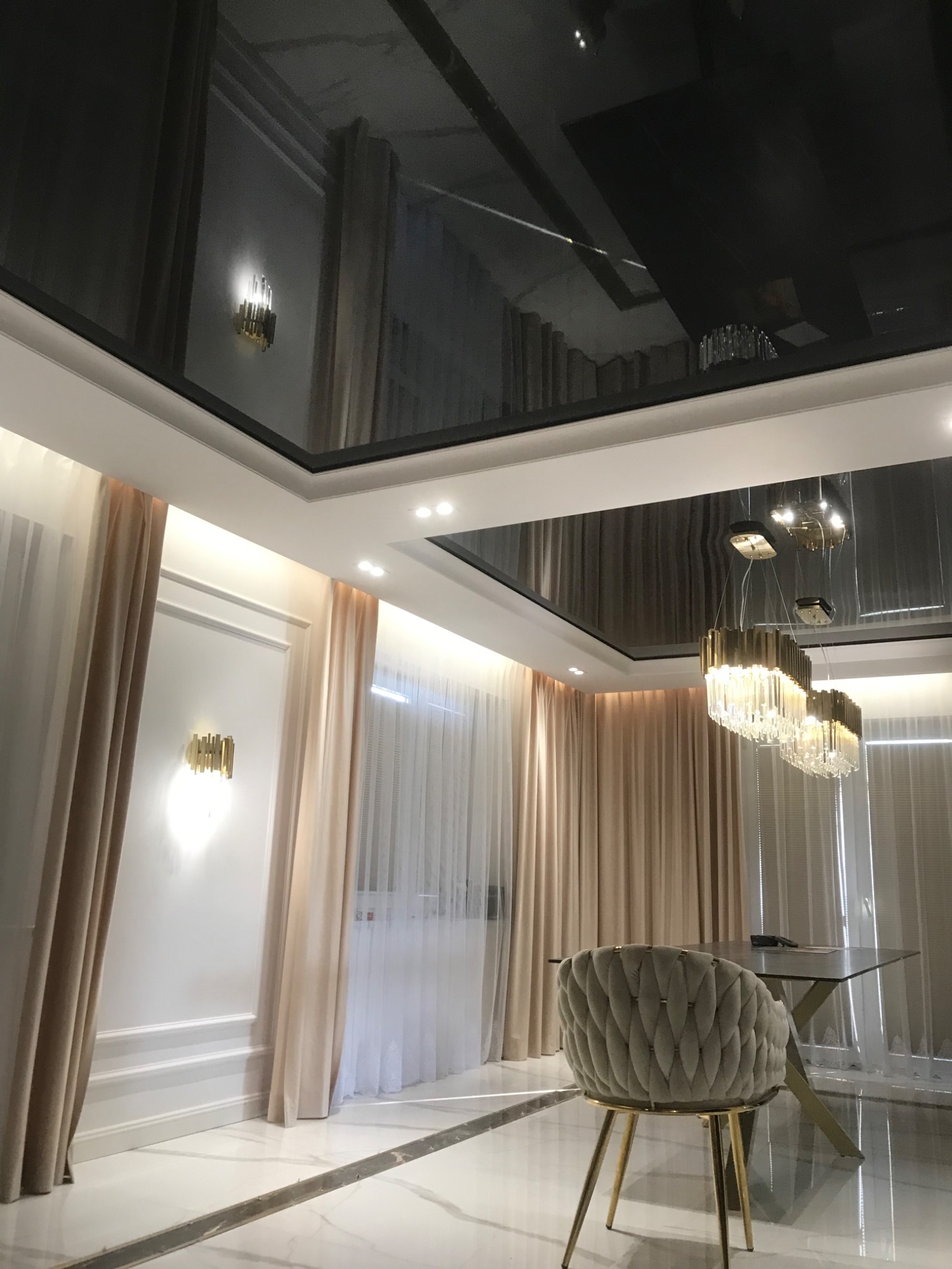 Nowoczesne wnętrze z czarnym, błyszczącym sufitem napinanym, odbijającym kryształowy żyrandol i złote kinkiety, jasne zasłony, białe ściany z dekoracyjnymi listwami i marmurowa podłoga.