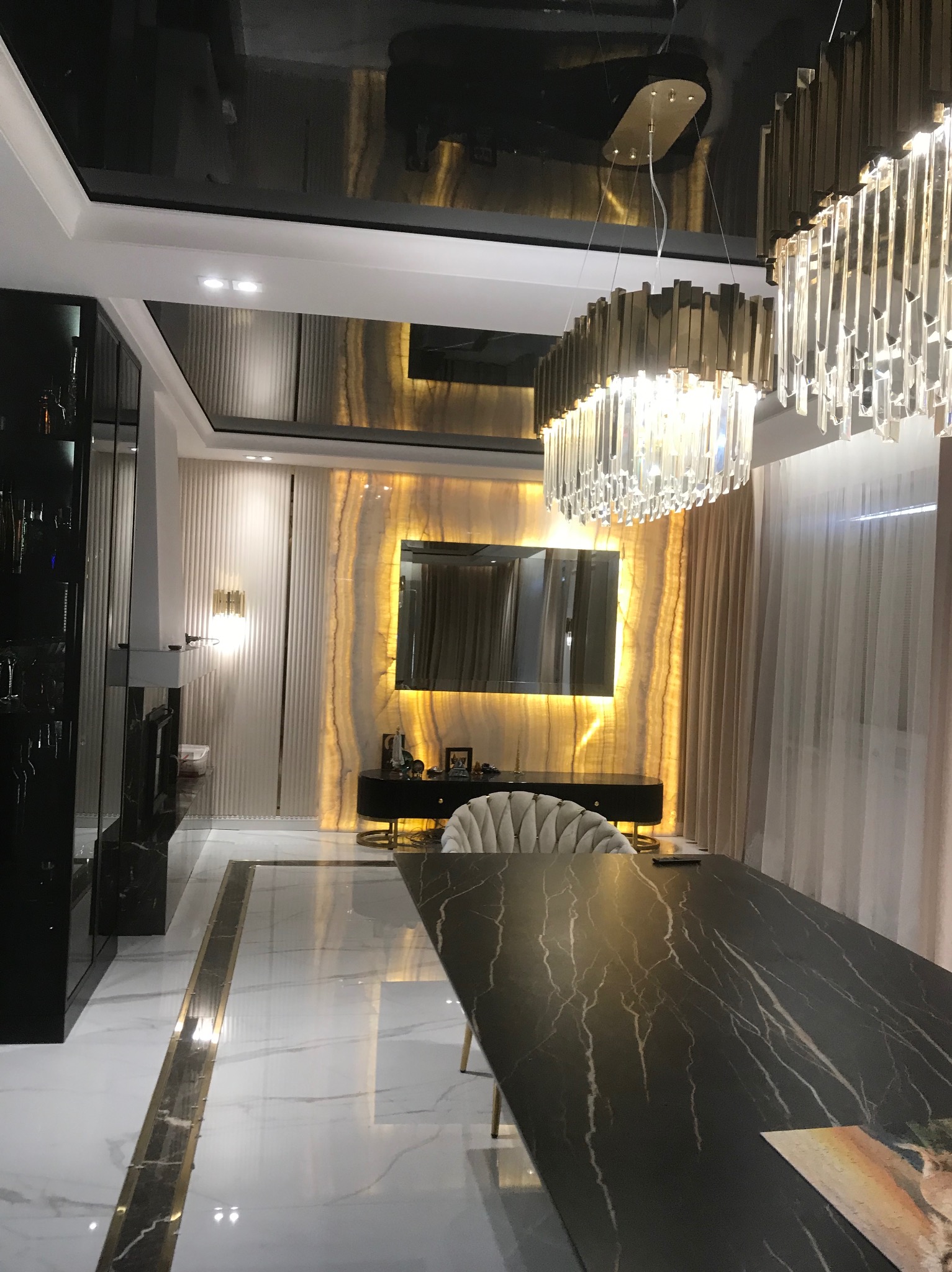 Eleganckie wnętrze jadalni z czarnym, błyszczącym sufitem, marmurową podłogą, podświetlaną ścianą z onyksu i kryształowym żyrandolem nad ciemnym stołem.