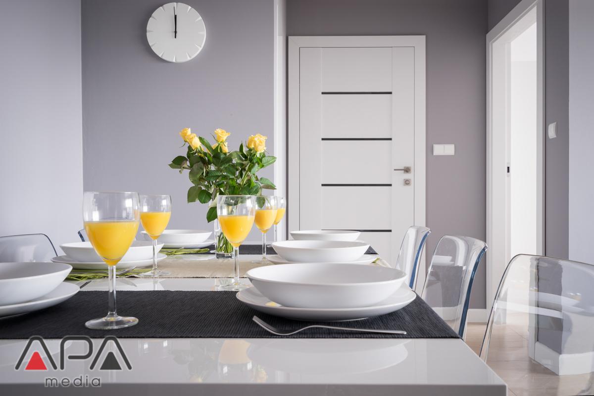 Nowoczesna jadalnia z białym stołem, nakrytym dla kilku osób, z żółtymi różami w wazonie i szklankami z sokiem pomarańczowym, na tle szarej ściany z białym zegarem.