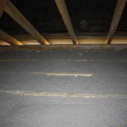 Poddasze z widoczną konstrukcją drewnianą i szarą warstwą izolacyjną pomiędzy belkami, wykonaną z materiału włóknistego.