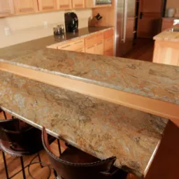 Kuchnia z blatem o wzorze przypominającym piaskowiec, oddzielona od części jadalnej podwyższonym barem z trzema krzesłami barowymi z ciemnej skóry.