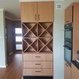 Drewniany regał kuchenny z szafką, szufladami i półkami na wino, z jedną butelką wina, wkomponowany w zabudowę kuchenną z piekarnikiem.