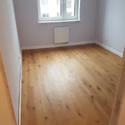 Pokój z nową, jasną podłogą drewnianą ułożoną w jodełkę, widok zza surowej ościeżnicy, ściany pomalowane na jasny szary kolor, biały grzejnik pod oknem.