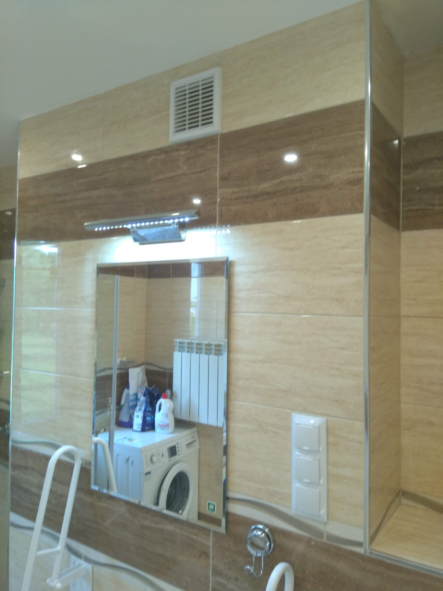 Beżowo-brązowa łazienka z lustrem, w którym odbija się pralka i środki czystości, widoczna kratka wentylacyjna i potrójny włącznik.