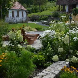 Kaskadowy ogród z drewnianym tarasem i leżakami na tle dwóch budynków z czerwonymi dachami, ścieżka z drobnego żwiru.