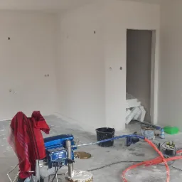 Agregat malarski SmartControl na kółkach z czerwoną szmatką, wiadro z farbą i akcesoria malarskie w surowym, pomalowanym na biało pomieszczeniu w trakcie prac malarskich.