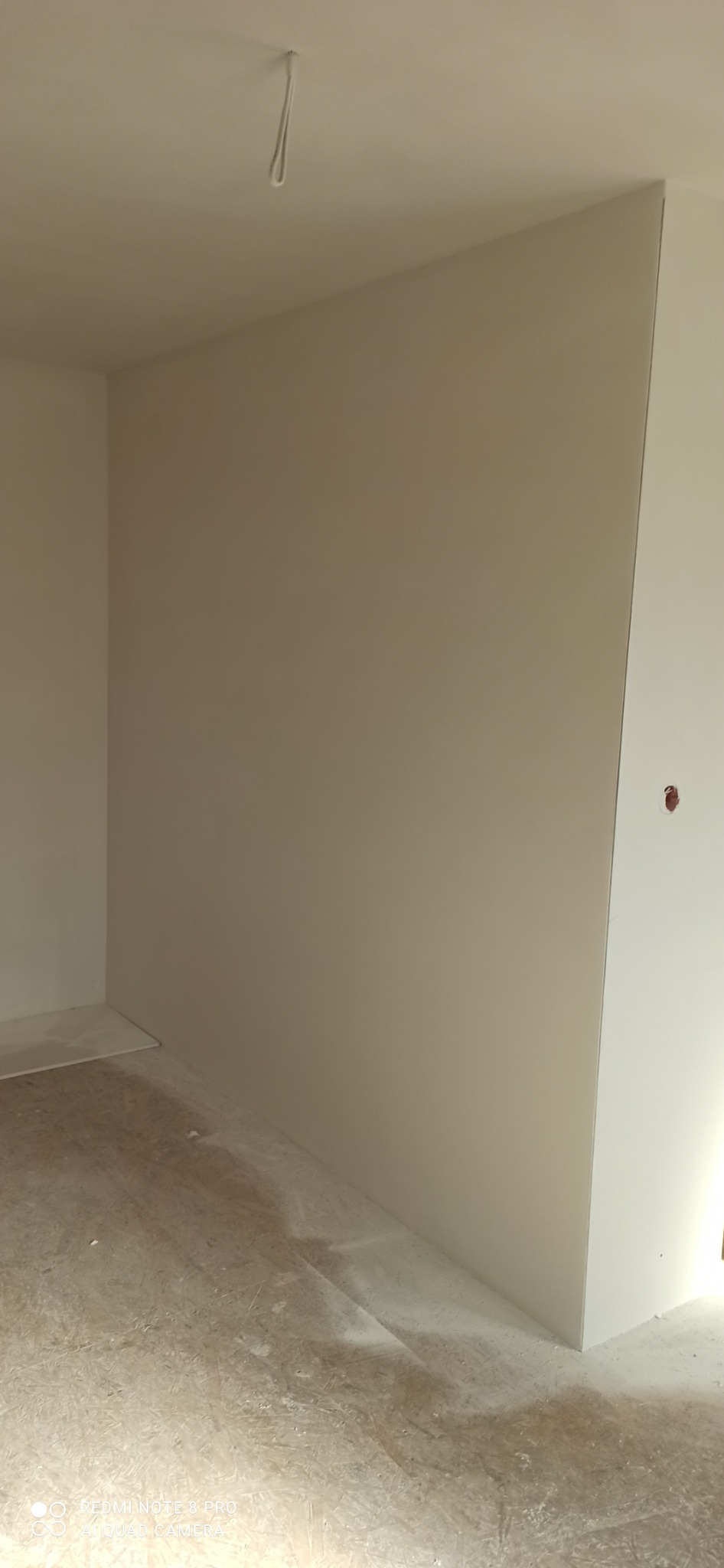 Świeżo wykończony narożnik pokoju z gładką, beżową ścianą i białą płytą kartonowo-gipsową, widoczne ślady po pracach wykończeniowych na podłodze, sufit z wystającym elementem mocującym.