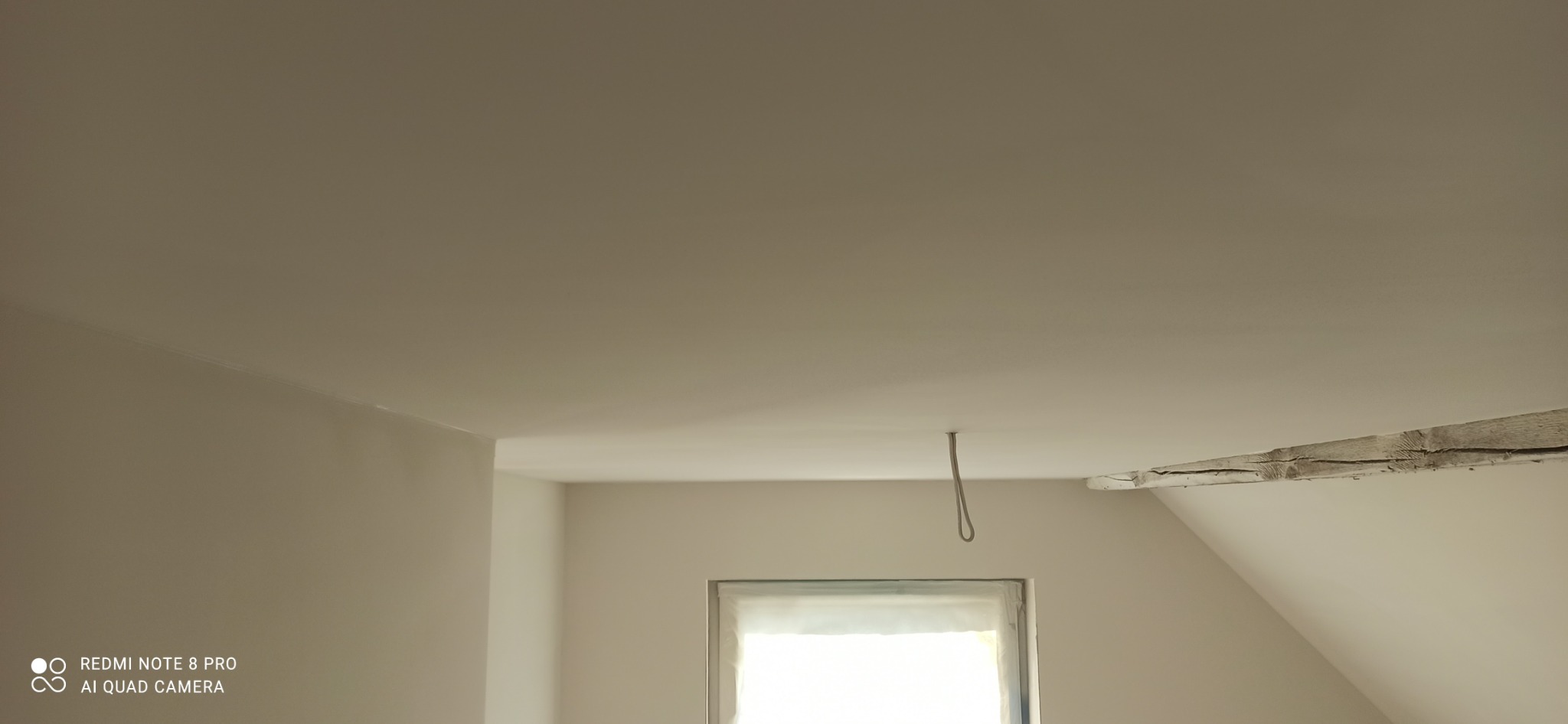 Wnętrze poddasza po malowaniu ścian na jasny odcień, widoczny fragment drewnianej belki stropowej i kabel wystający z sufitu, okno zabezpieczone folią malarską.