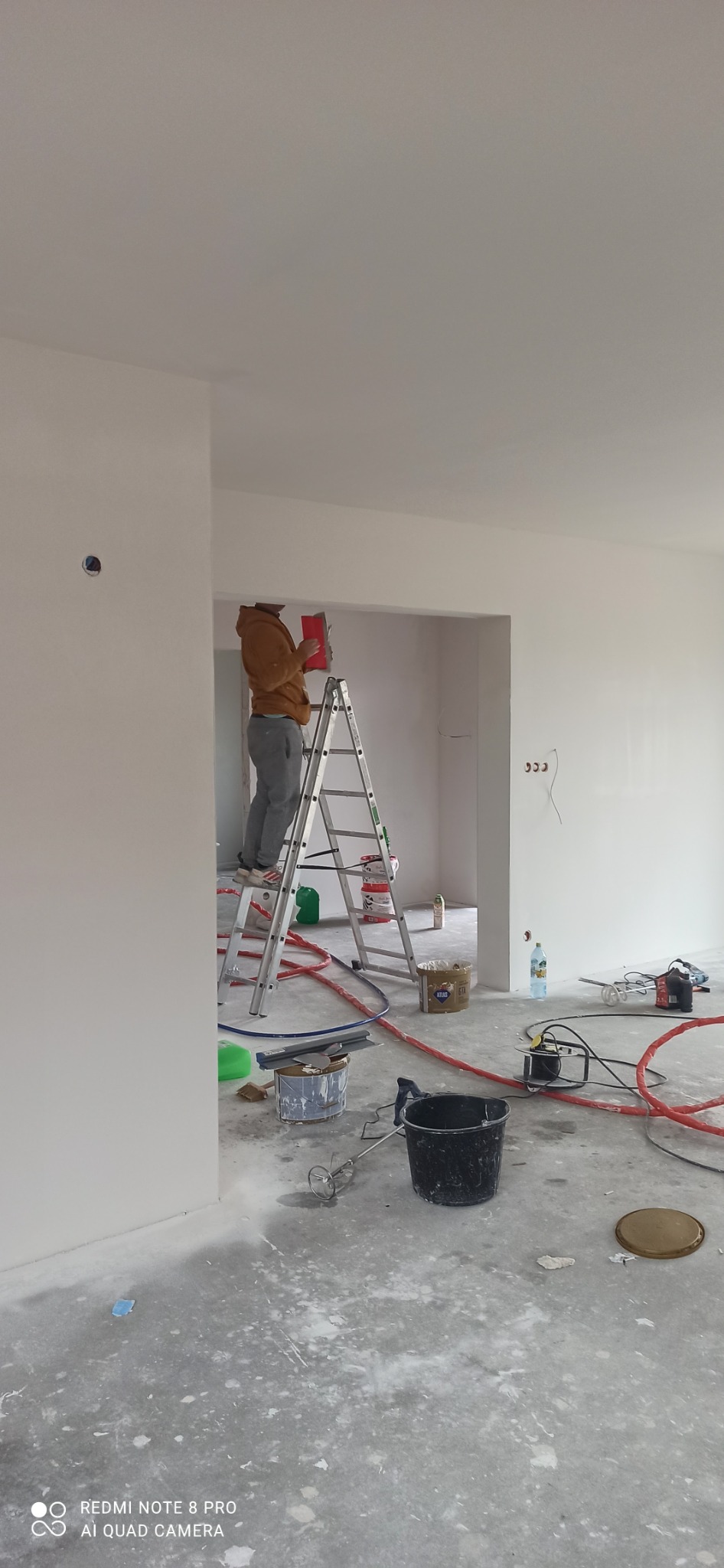 Pracownik na drabinie szpachluje ścianę w jasnym, pustym pokoju. Na podłodze widoczne narzędzia, wiadro, mieszadło i ślady po szpachlowaniu.