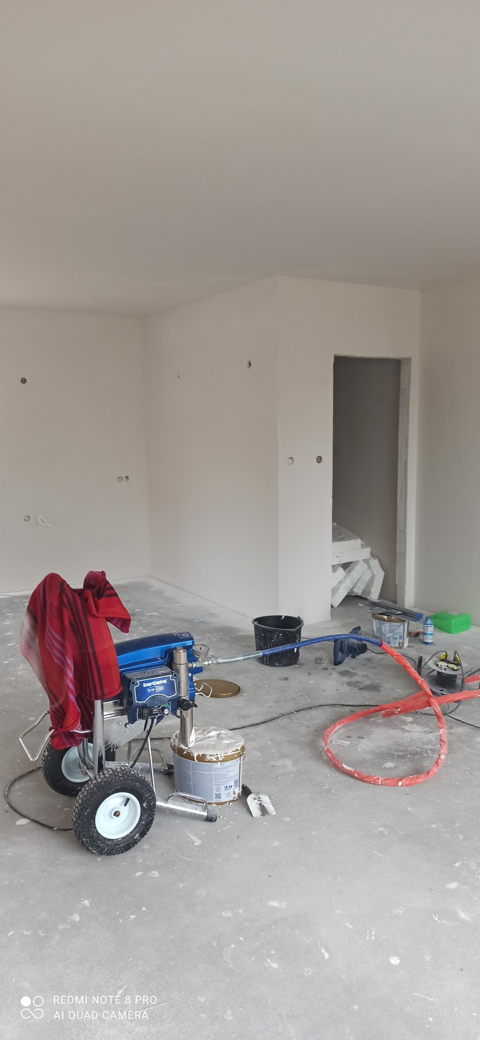 Agregat malarski SmartControl na kółkach z czerwoną szmatką, wiadro z farbą i akcesoria malarskie w surowym, pomalowanym na biało pomieszczeniu w trakcie prac malarskich.