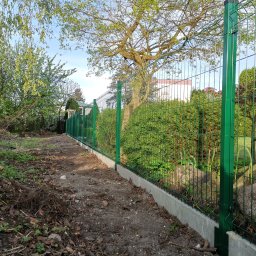 LIVBET-ECO - Ogrodzenia Systemowe - Zielone panele ogrodzeniowe z podmurówką betonową, widok z boku na tle drzew i krzewów w słoneczny dzień.