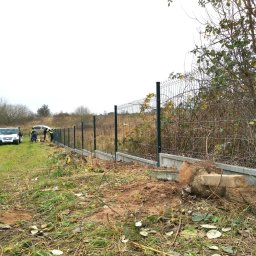 LIVBET-ECO - Ogrodzenia Systemowe - Montaż ogrodzenia panelowego z betonową podmurówką na nierównym terenie, widoczni pracownicy w odblaskowych kamizelkach i dwa samochody dostawcze w tle.