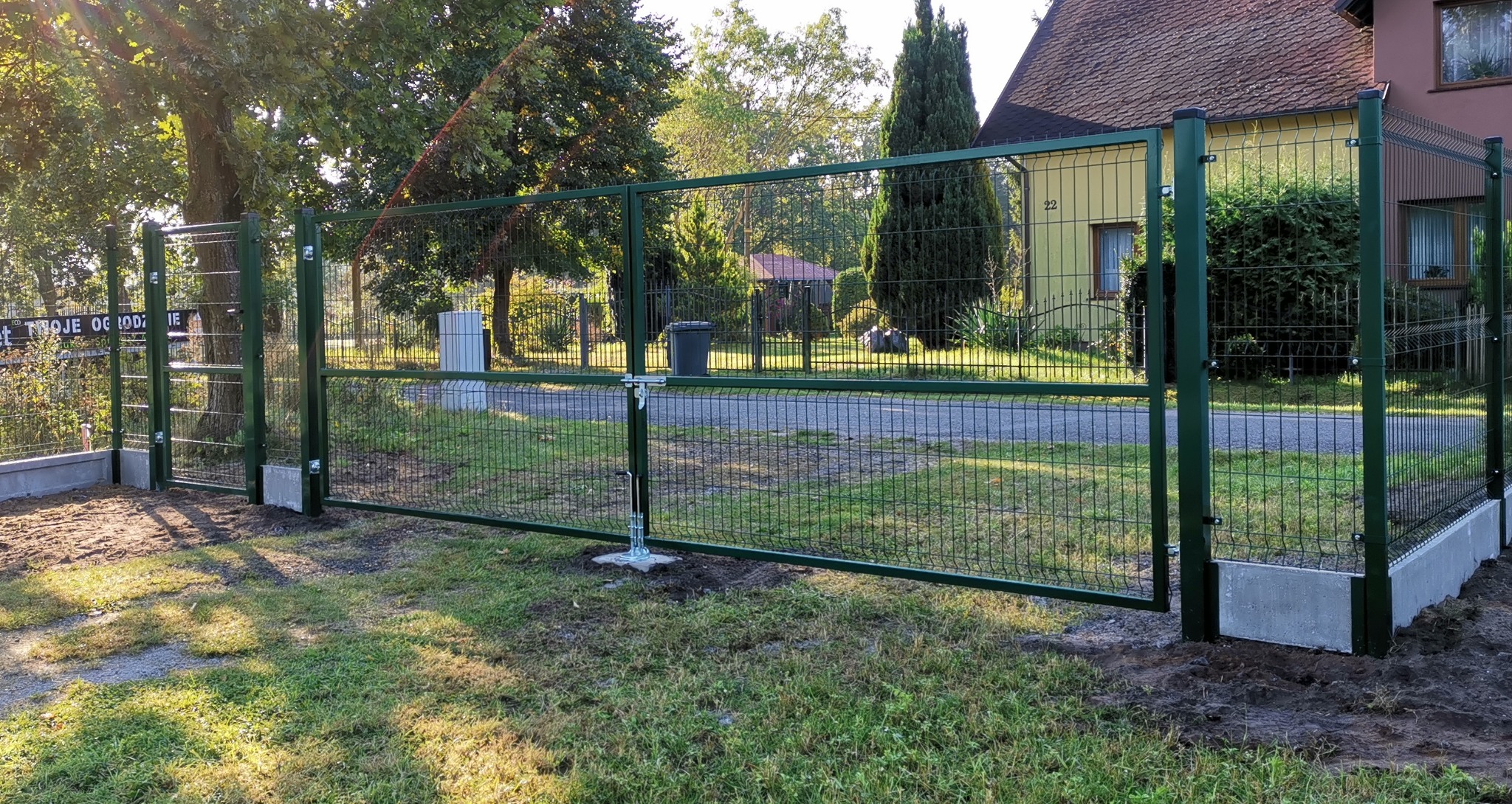 Zielona siatka ogrodzeniowa z furtką i bramą wjazdową, świeżo zamontowana na betonowej podmurówce, widok na dom i ulicę