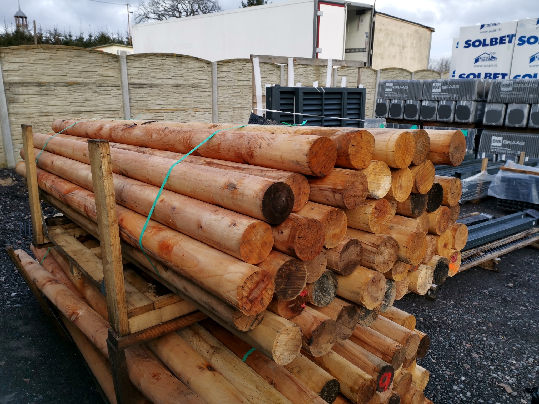 Stos impregnowanych drewnianych bali na palecie, przeznaczonych do budowy ogrodzeń, w tle betonowe panele ogrodzeniowe i materiały budowlane.