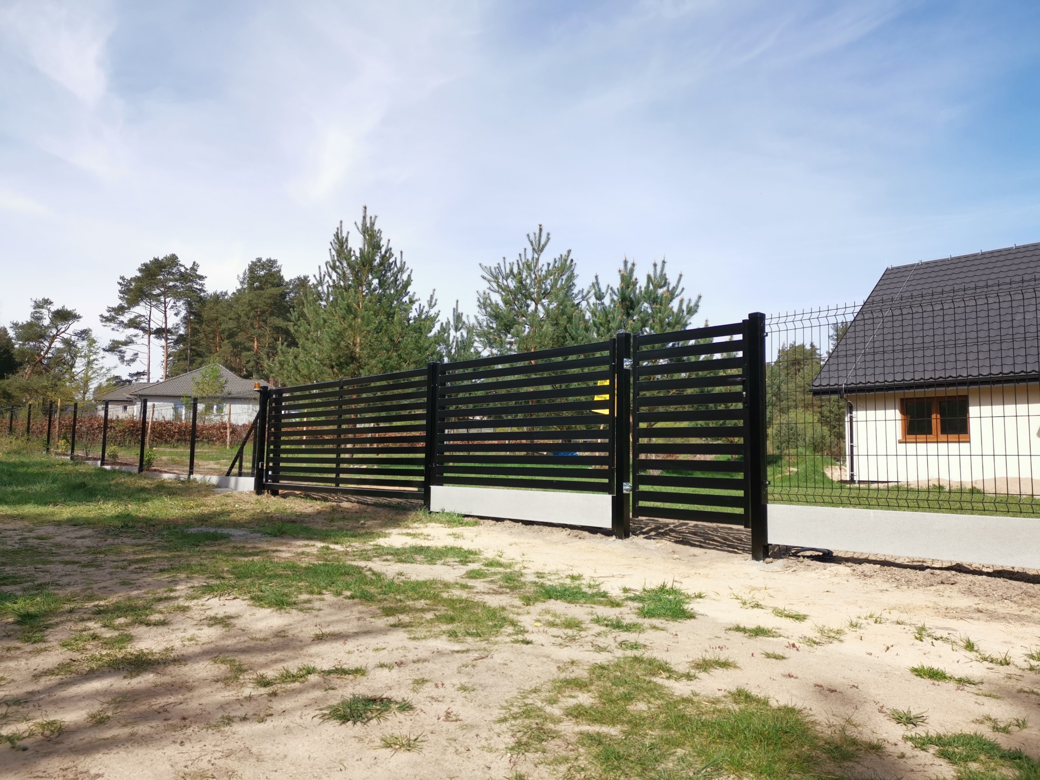 Nowoczesna, przesuwna brama wjazdowa z poziomych, czarnych paneli na betonowej podstawie, z widocznym fragmentem ogrodzenia panelowego i siatki ogrodzeniowej w tle, na piaszczystym terenie.
