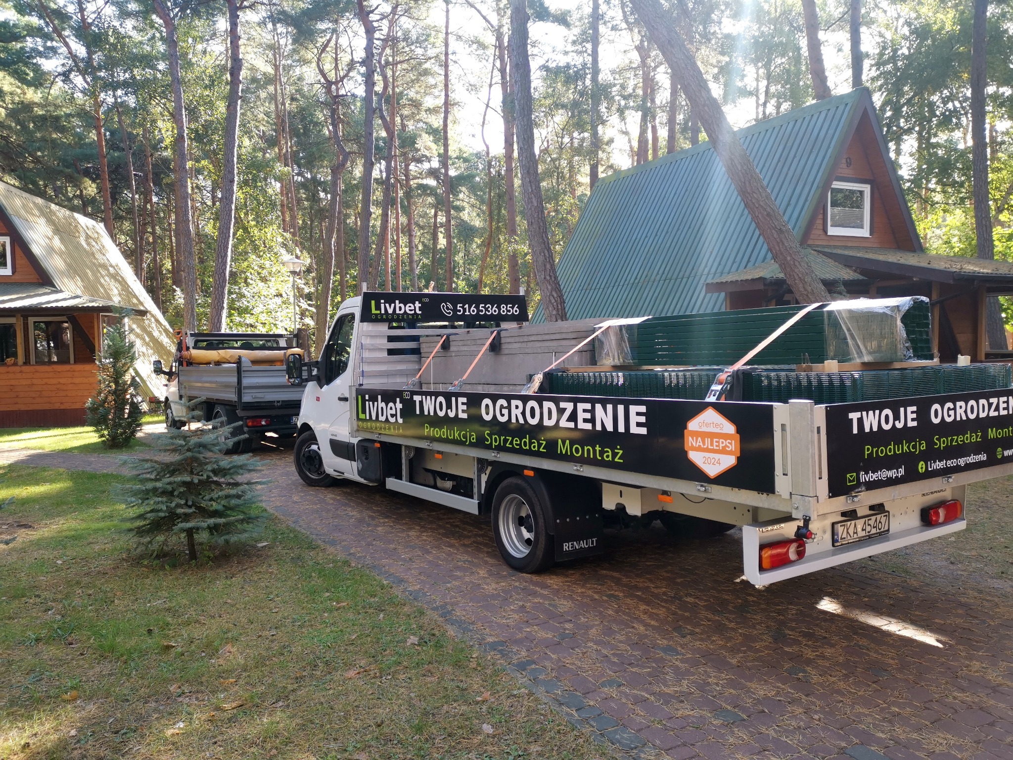Dwa samochody dostawcze firmy ogrodzeniowej na tle domków letniskowych i drzew, załadowane panelami ogrodzeniowymi i elementami montażowymi; na samochodach logo firmy i napis 'Twoje Ogrodzenie...