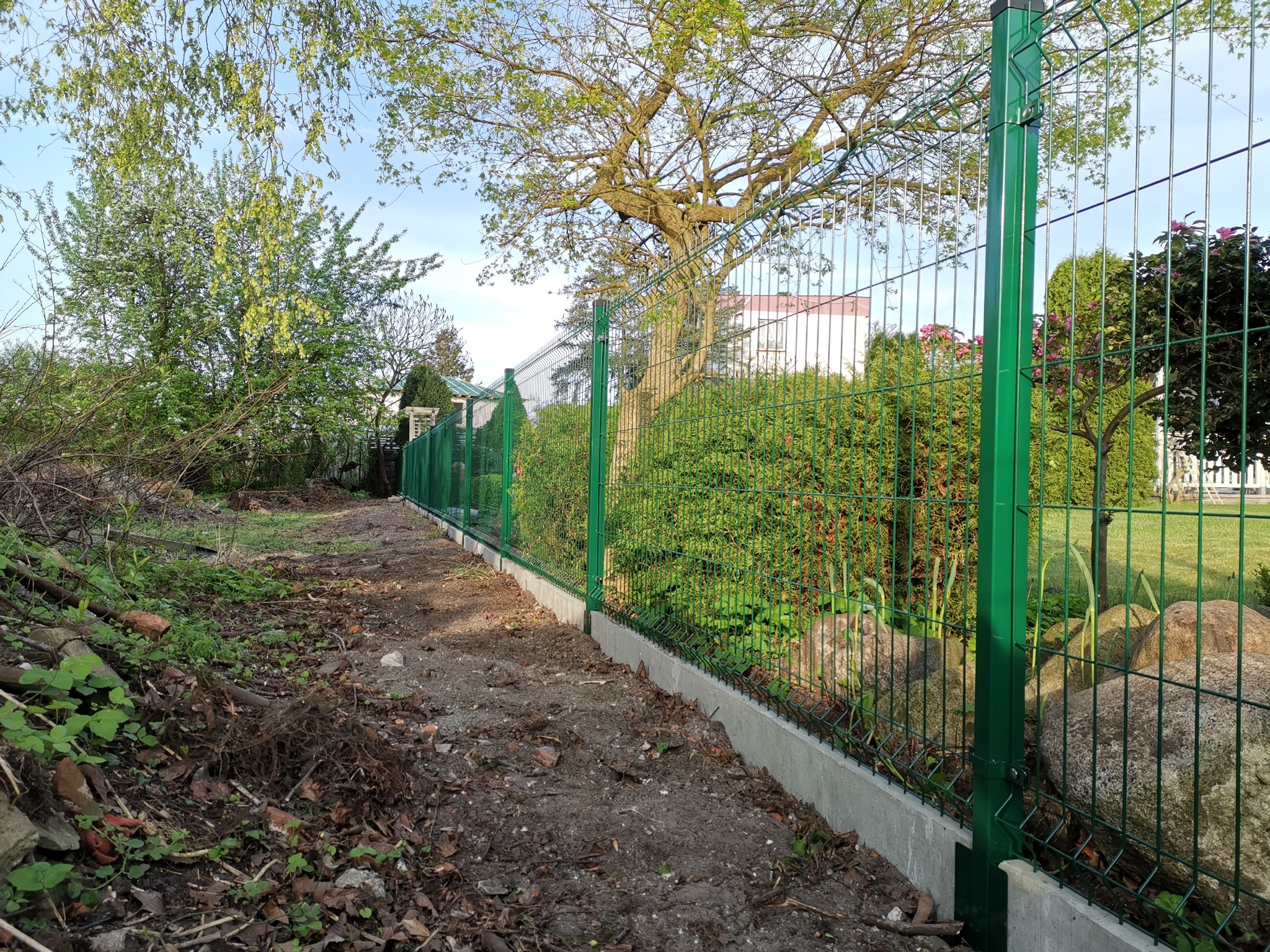 Zielone panele ogrodzeniowe z podmurówką betonową, widok z boku na tle drzew i krzewów w słoneczny dzień.