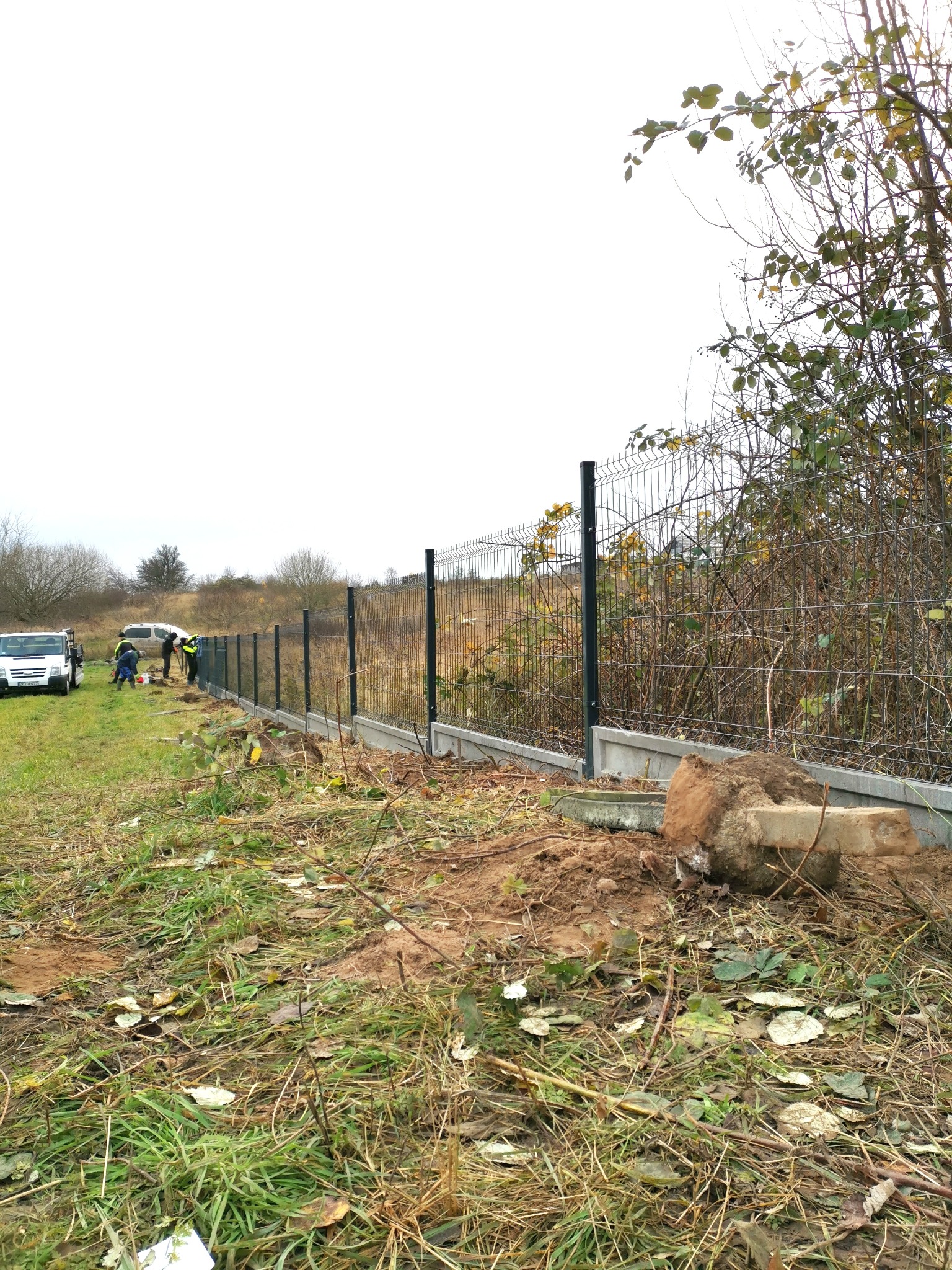 Montaż ogrodzenia panelowego z betonową podmurówką na nierównym terenie, widoczni pracownicy w odblaskowych kamizelkach i dwa samochody dostawcze w tle.