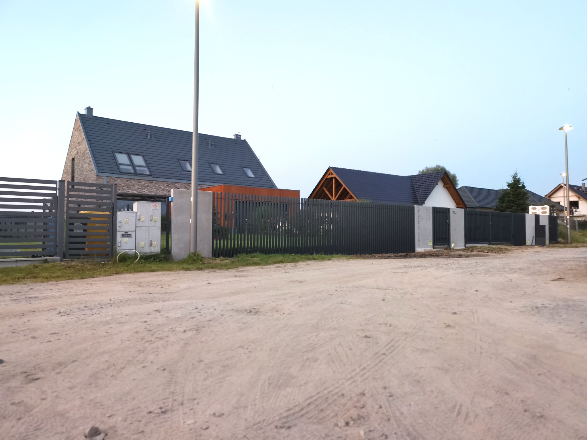 Nowoczesne, poziome ogrodzenie panelowe w kolorze grafitowym, z betonowymi słupkami, na tle nowo wybudowanych domów jednorodzinnych i piaszczystej drogi.