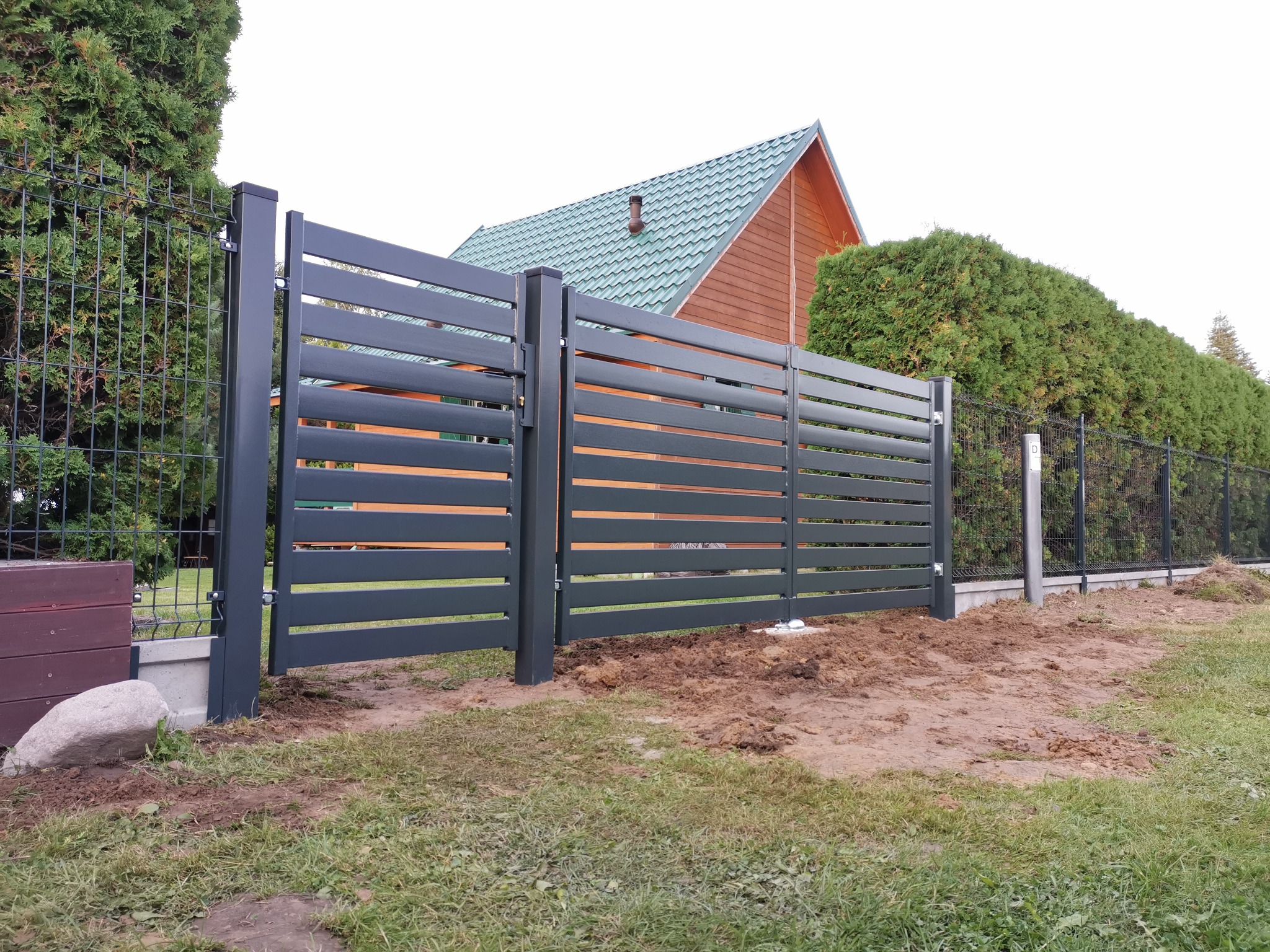 Nowoczesna brama wjazdowa z poziomych paneli w kolorze grafitowym, zamontowana przy ogrodzeniu z siatki, widoczny dom z zielonym dachem w tle.