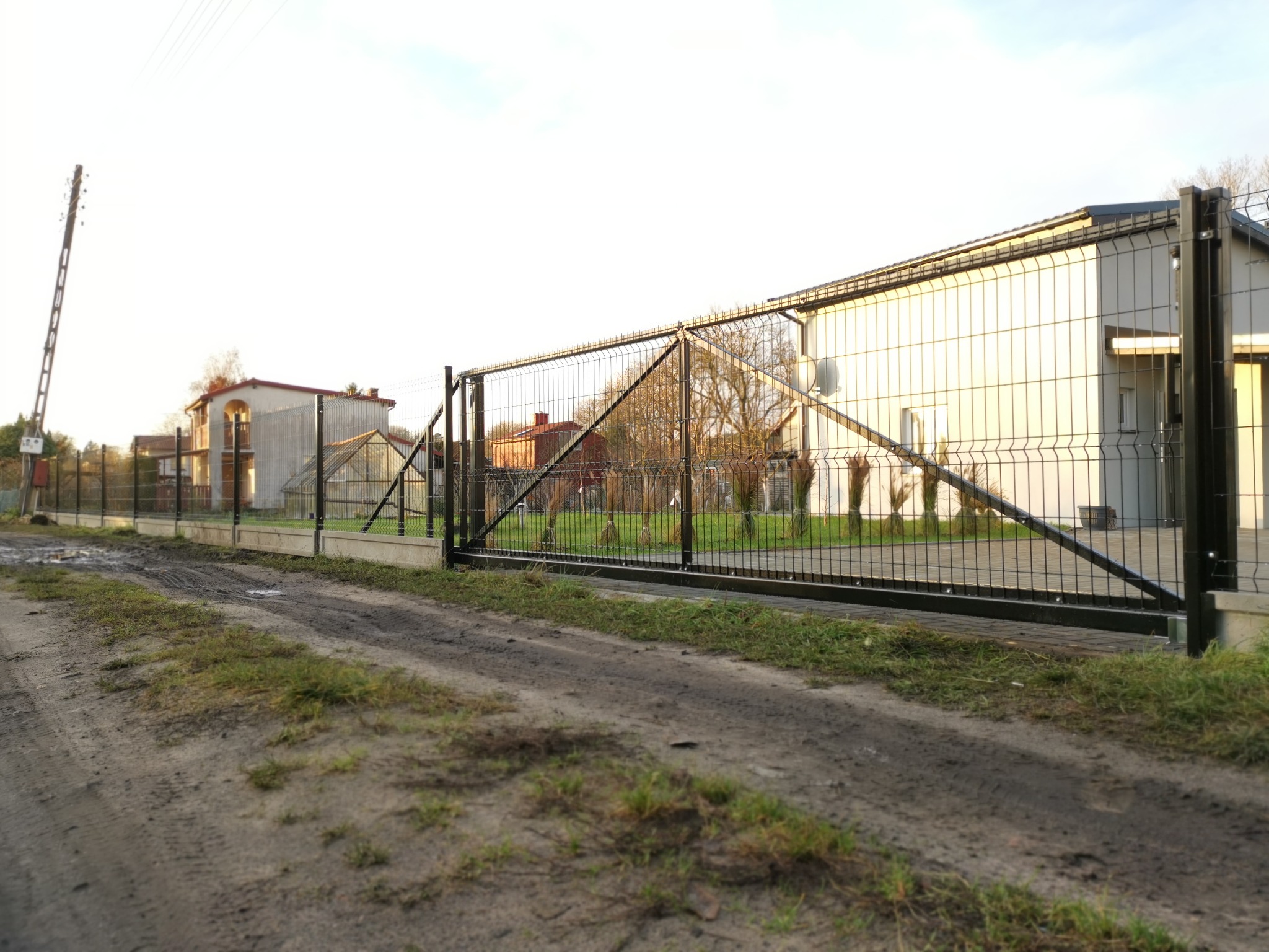 Czarna brama wjazdowa z siatki stalowej, widok z polnej drogi, w tle domy jednorodzinne i słup energetyczny.