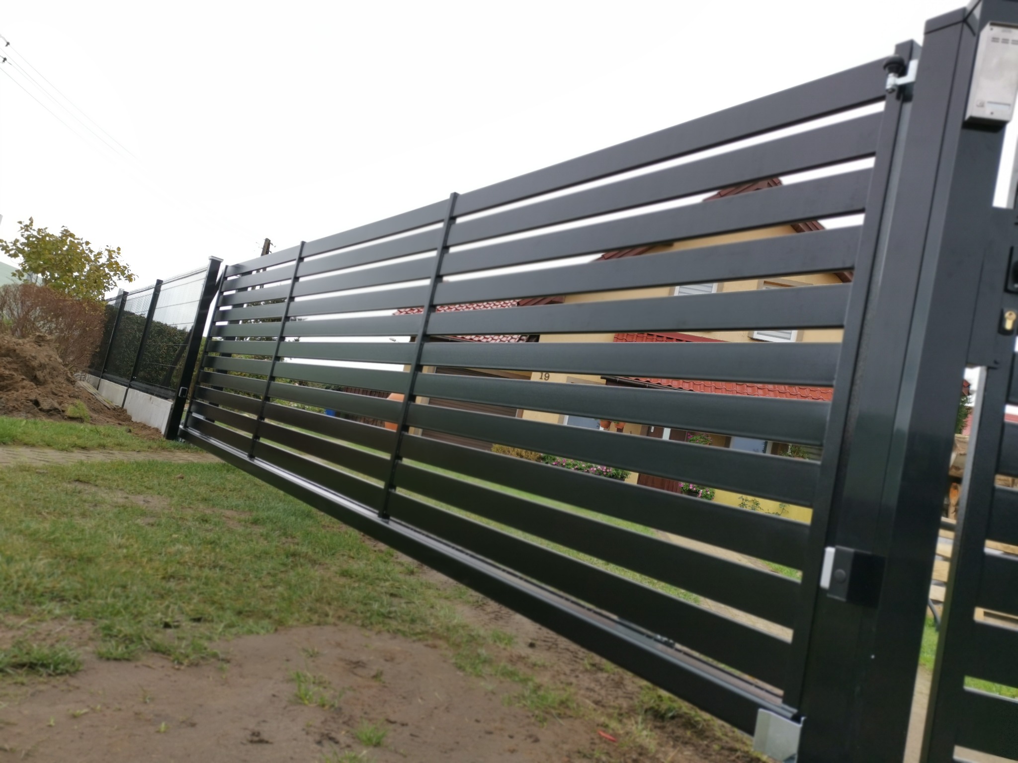 Nowoczesna, pozioma brama wjazdowa z czarnych paneli, widoczny zamek i dom w tle, trawnik po deszczu.