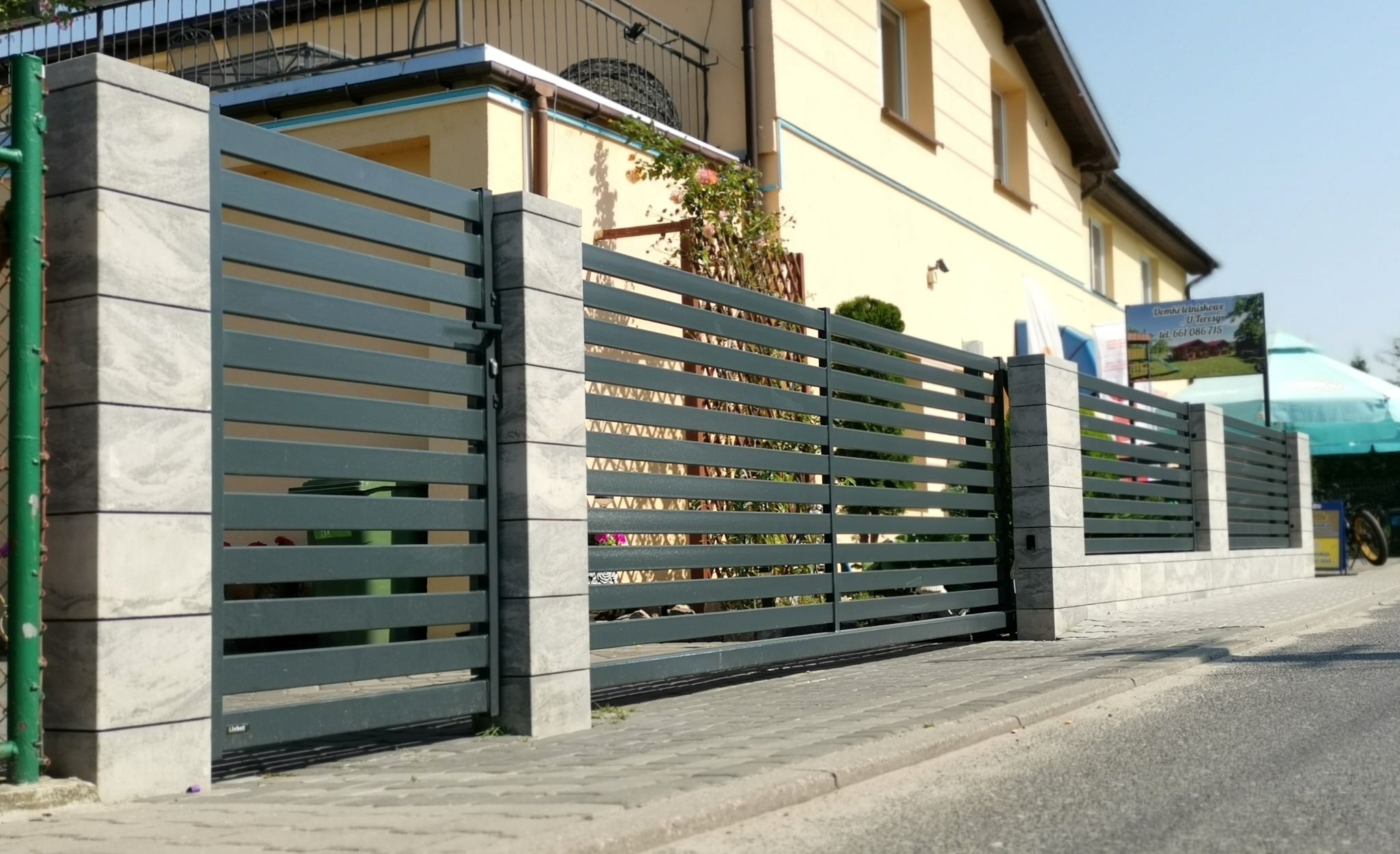 Nowoczesna brama wjazdowa z szarymi poziomymi panelami i kamiennymi słupkami, widok z poziomu ulicy na tle domu i reklamy.